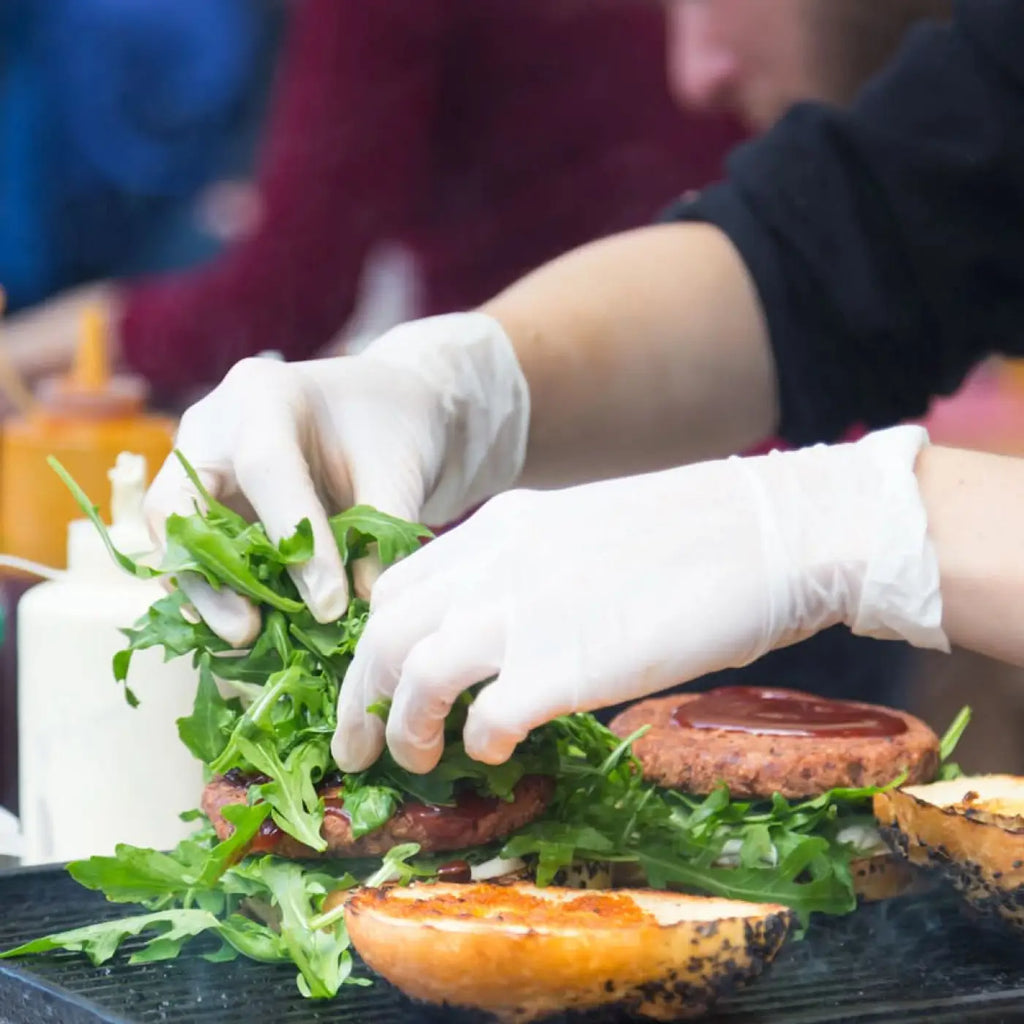 Die hände eines kochs in weißen hygienischen nitrilhandschuhen von hygostar platzieren frischen rucola auf einem burger mit grillspuren