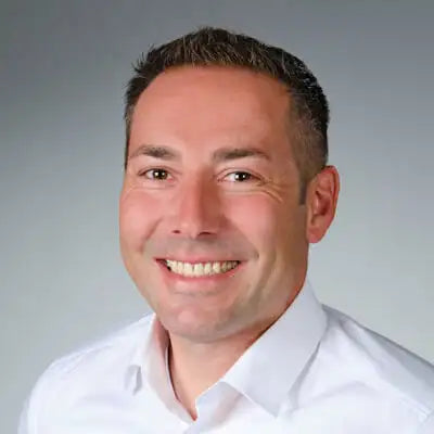 Lächelnder Mann mit kurzem Haar trägt weißes Hemd vor neutralem grauem Hintergrund