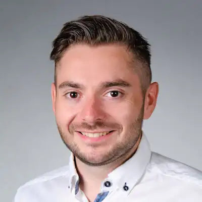 Porträt eines lächelnden Mannes mit kurzem Haar und weißem Hemd vor grauem Hintergrund.