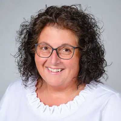 Porträt einer lächelnden Frau mit lockigen dunklen Haaren und Brille vor neutralem Hintergrund