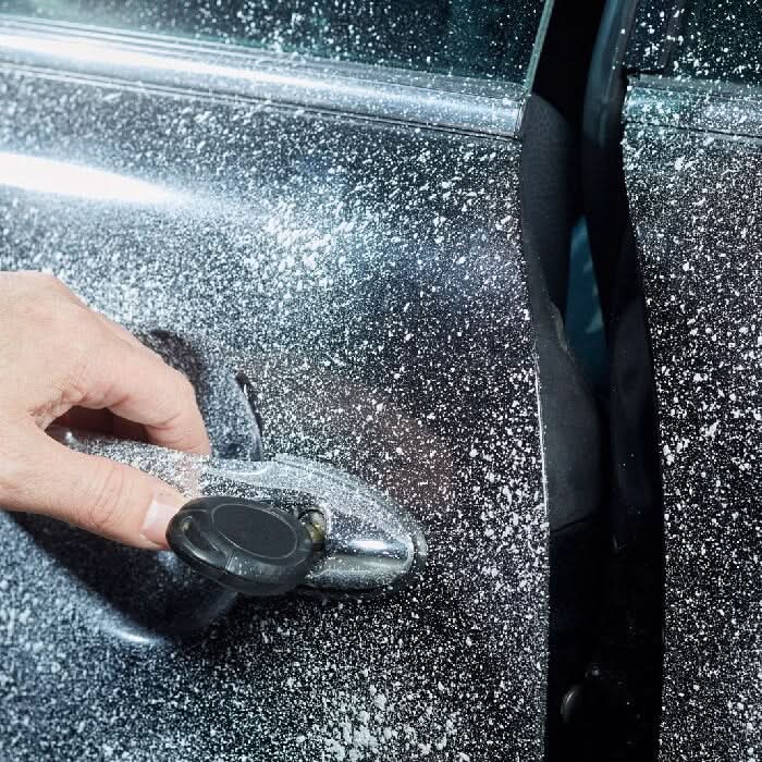 Eine hand hält einen schwarzen autotuerschloss mit einem silberschlüssel vor einem dunklen autospritzlack mit sonax schlossenteiser 50ml