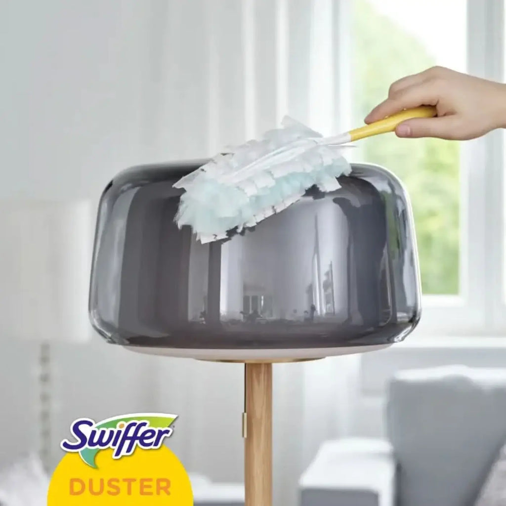 Swiffer xxl duster mit gelbem griff und weißen mikrofasertüchern reinigt obere fläche einer grauen glasklampe