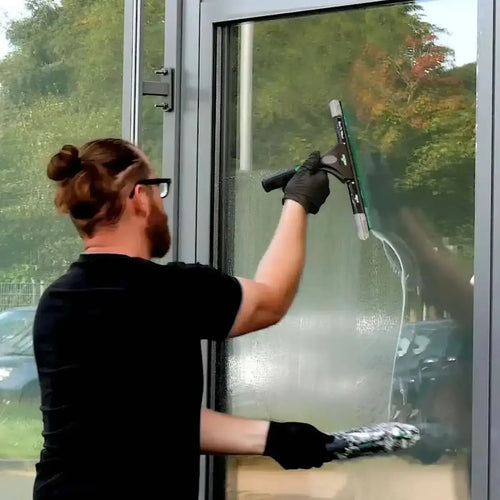 Schwarzhändige hand mit gummihandschuh hält unger ergotec ninja fensterwischer mit grünem gummiblatt und schwarzem griff zur glasreinigung